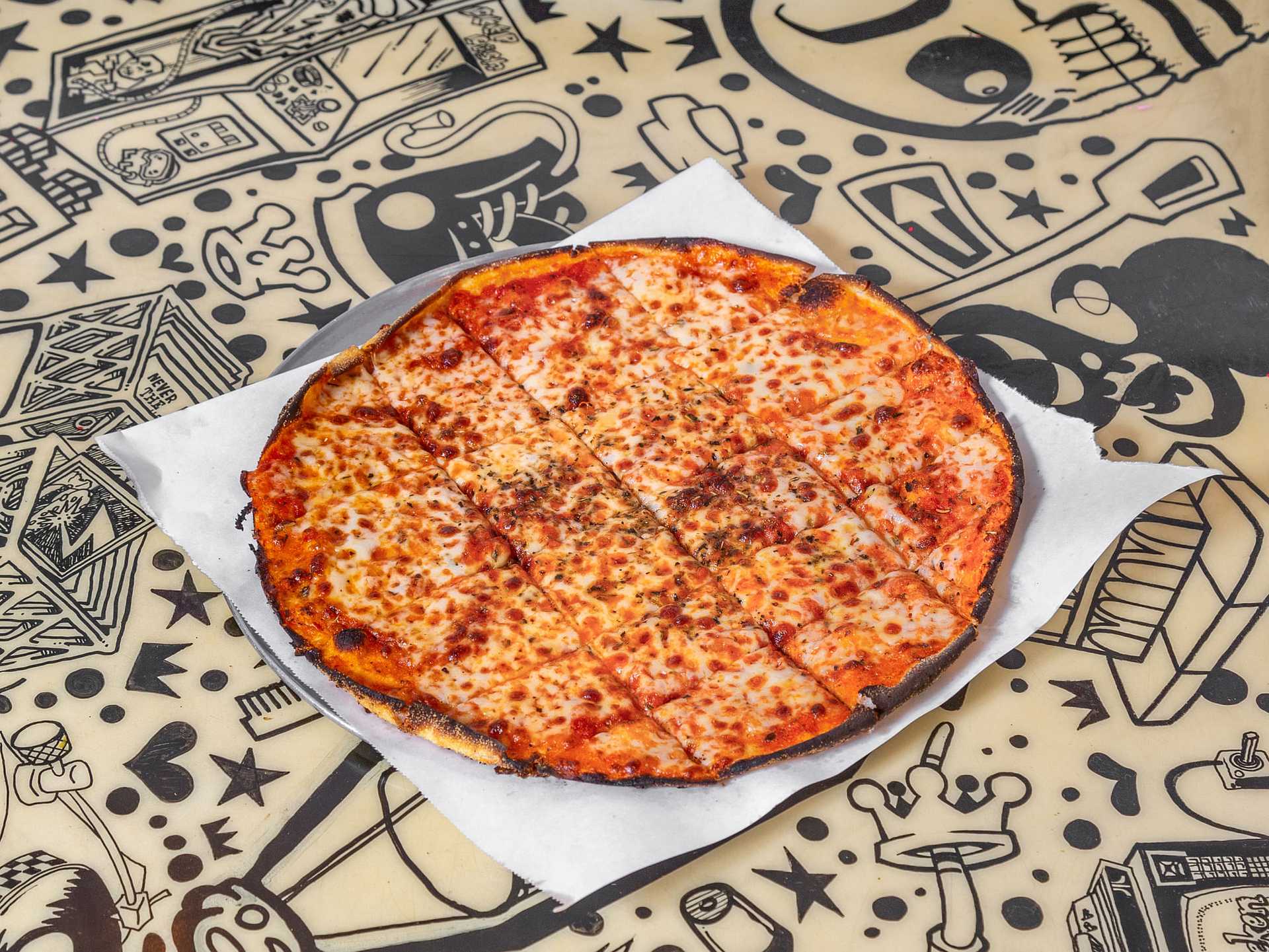 Thin-crust cheese pizza on white parchment paper, placed on a doodle-patterned table.