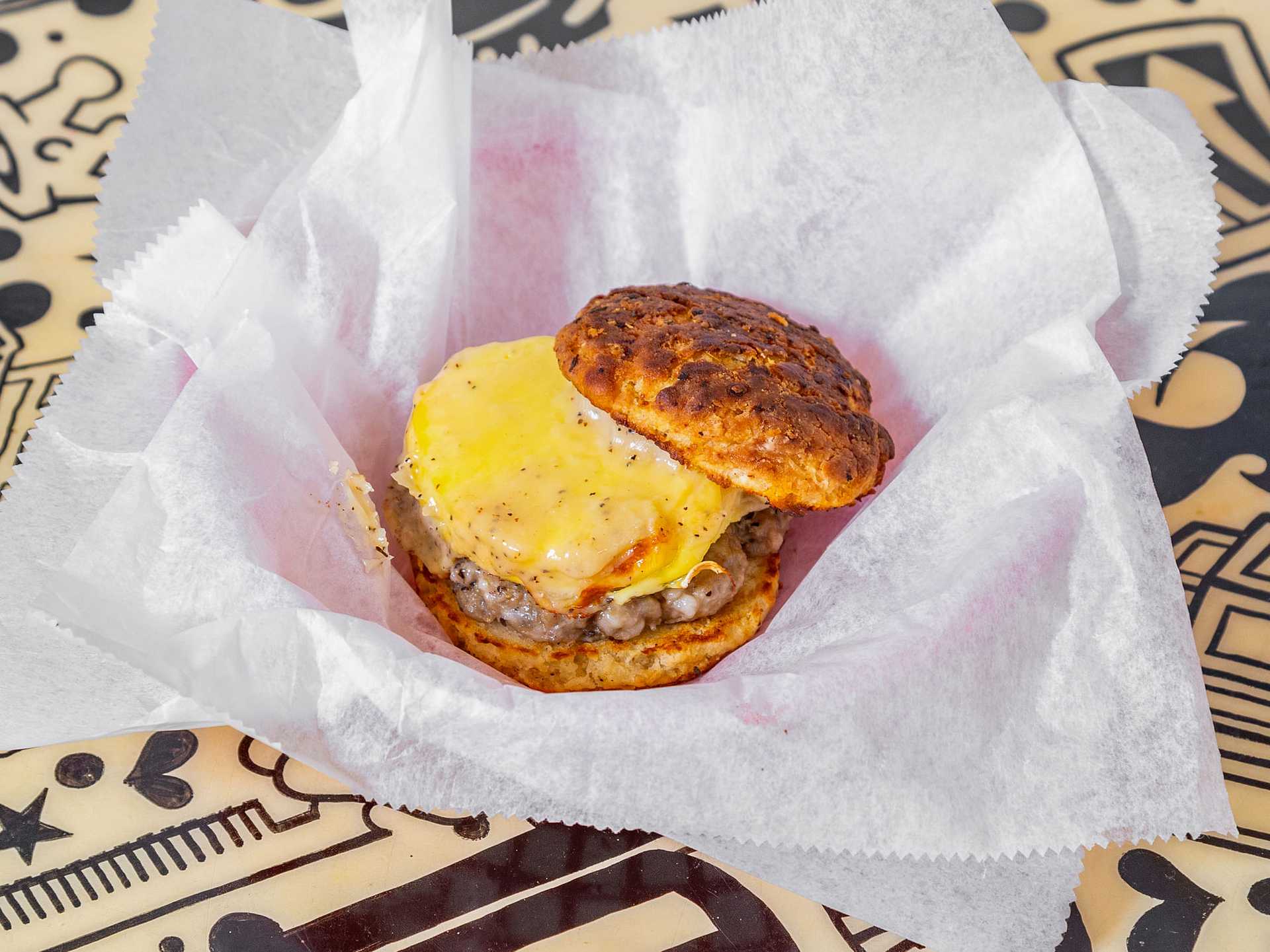 Cheeseburger with egg on a biscuit, wrapped in white paper on a patterned table.
