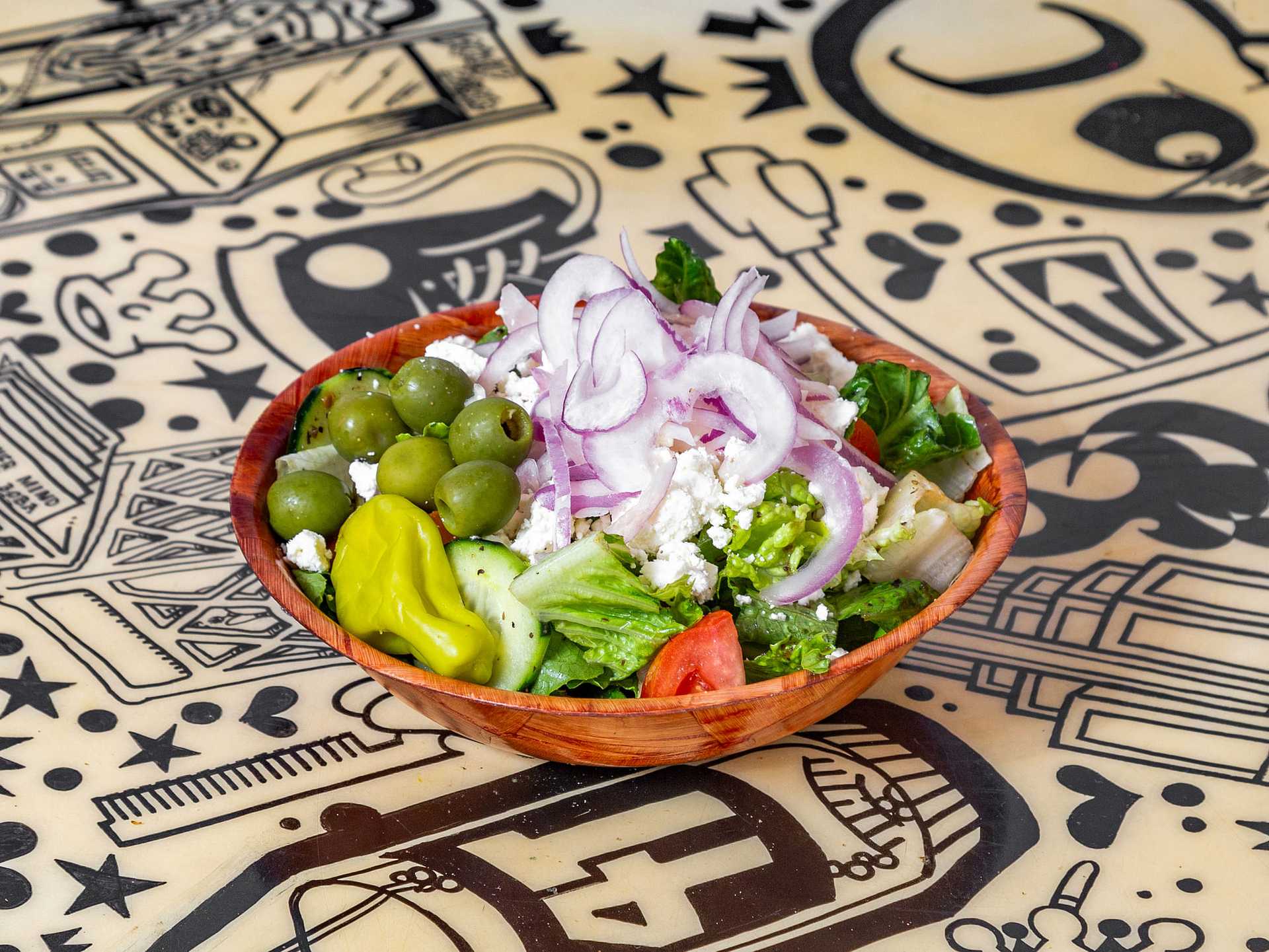 Salad bowl with olives, feta cheese, red onions, tomatoes, and greens on a patterned table.